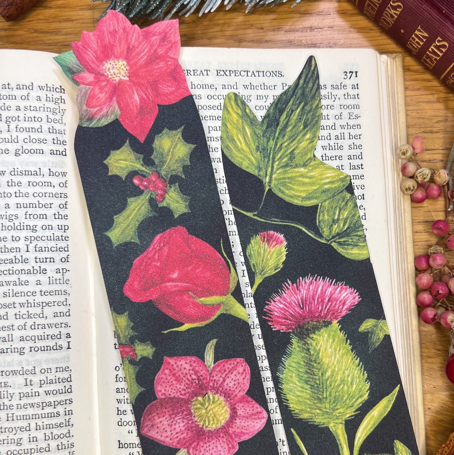 2 winters bloom bookmarks one decorated with illustrated rose, hellebore, holly and poinsettia, the other with thistle and ivy, both sat in the open pages of a book