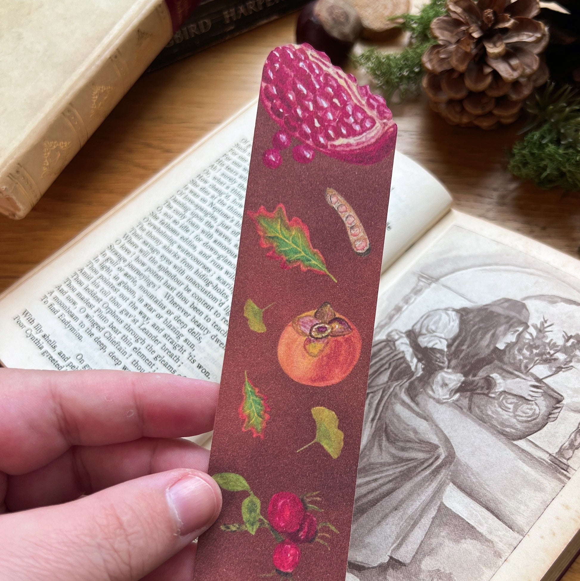 Woodland Walk Fruits Bookmark held in front of an open book. Bookmark features a pomegranate cut out at the top, and has autumnal leaves, persimmon, rose hips and seed pods illustrated across a warm toned brown background.