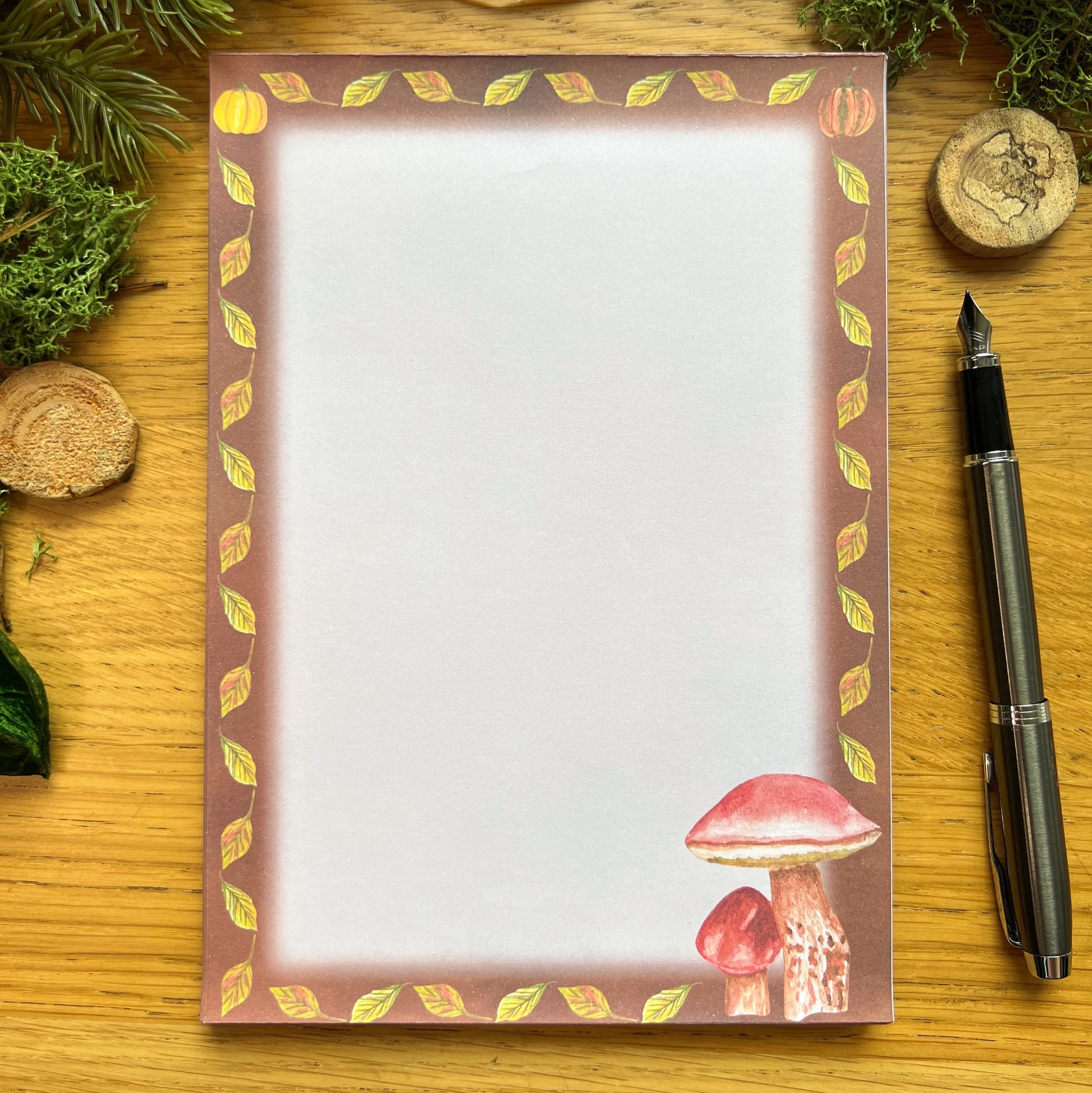 Autumnal mushroom illustrated notepad with warm brown leaf and pumpkin illustrated border around the page. The writing paper is blank. Notepad is on a wooden desk decorated with green moss, straw, pine cones and wooden slices and a fountain ink pen