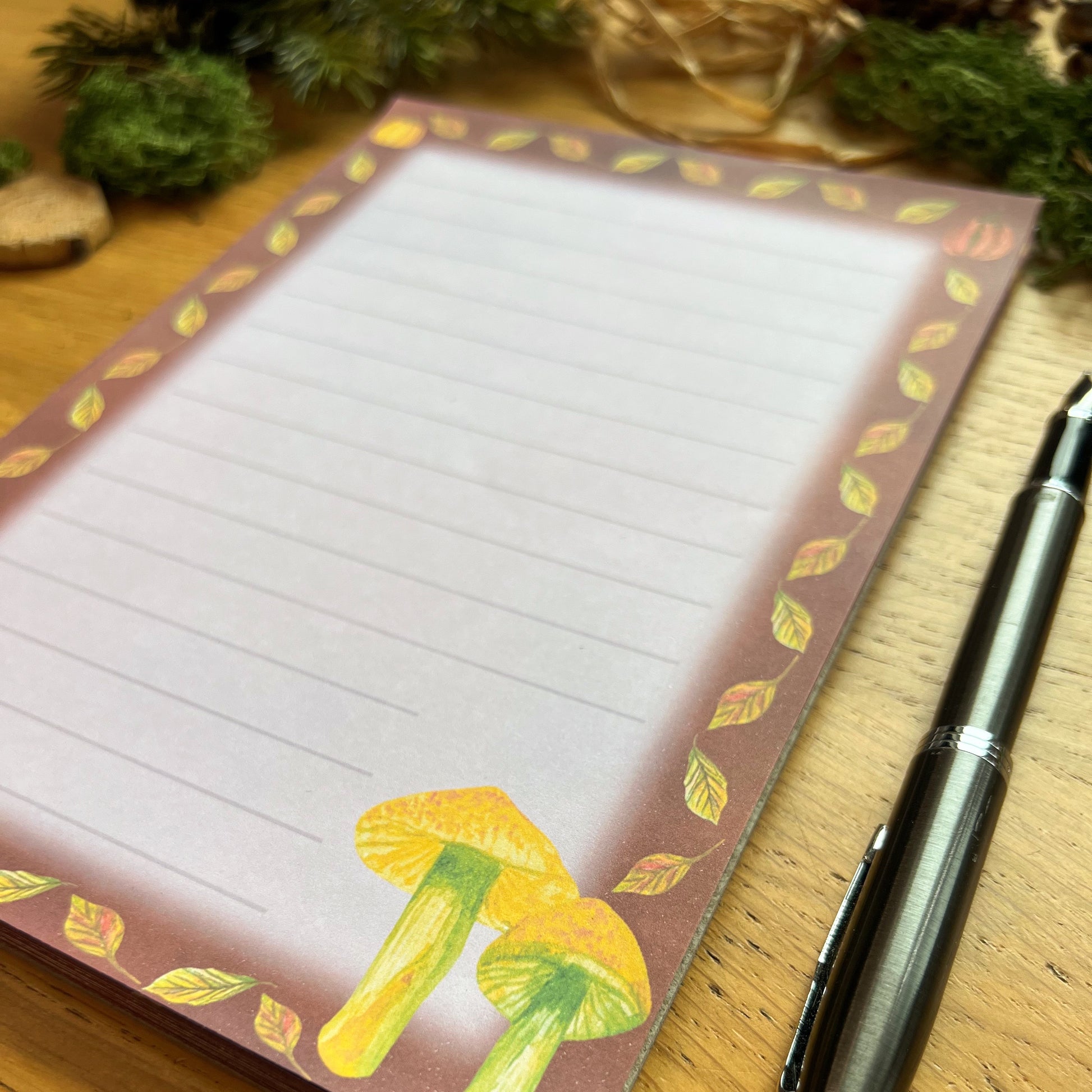 Autumnal mushroom illustrated notepad with warm brown leaf and pumpkin illustrated border around the page. The writing paper is subtly lined. Notepad is on a wooden desk decorated with green moss, straw, pine cones and wooden slices and a fountain ink pen