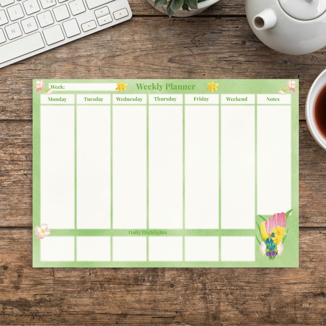 Spring floral botanical weekly planner with light green background, illustrated with cherry blossoms, daffodils and a spring bouquet.  White column sections for each day of the week have smaller daily highlights sections underneath each day, and to the left is a notes column for writing in also.  The pad sits on a wooden desk next to a cup of tea and a keyboard.