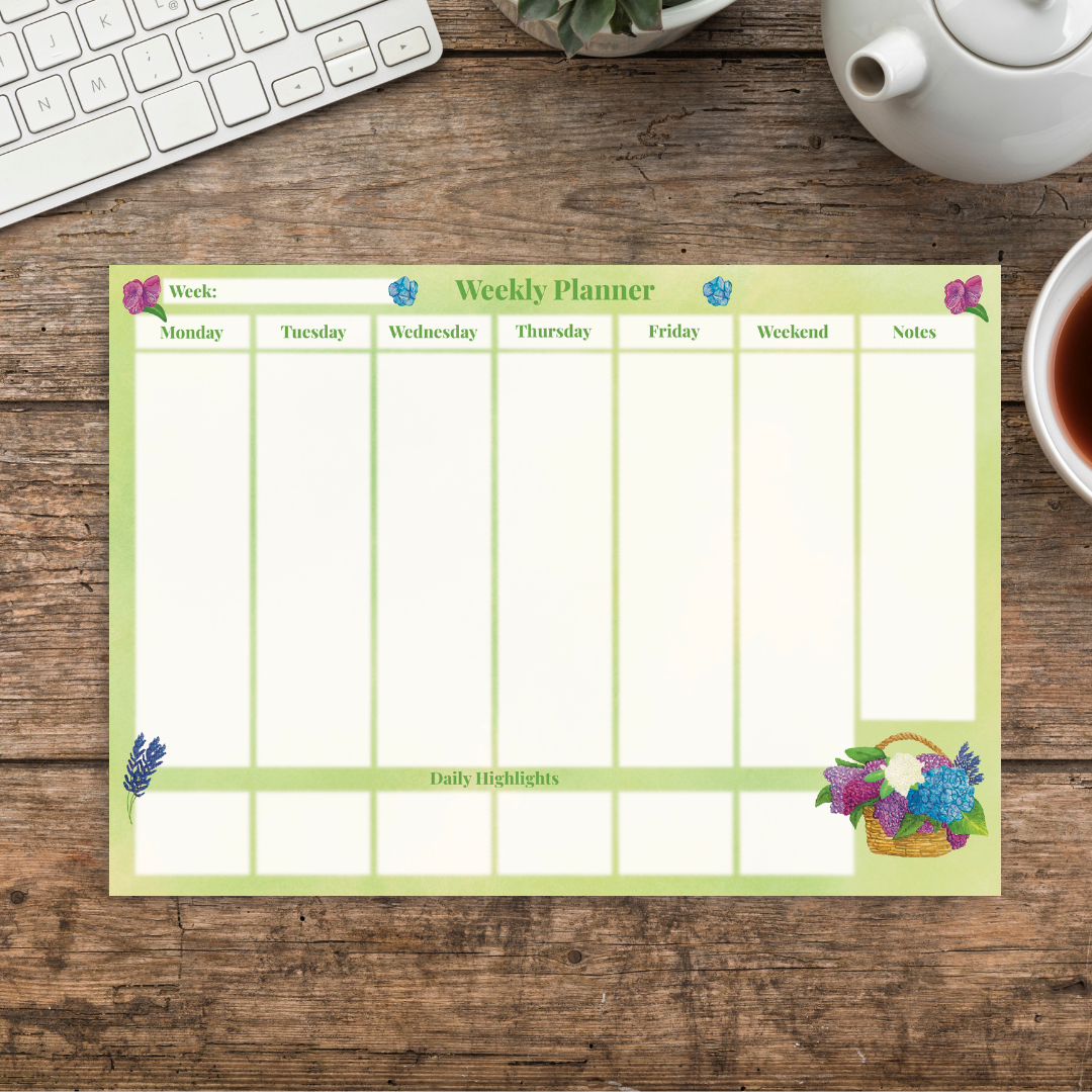 Summer botanical weekly planner with bright green background, illustrated with magenta, blue and purple florals, with a basket of lilacs on the bottom right corner.  White column sections for each day of the week have smaller daily highlights sections underneath each day, and to the left is a notes column for writing in also.  The pad sits on a wooden desk next to a cup of tea and a keyboard.