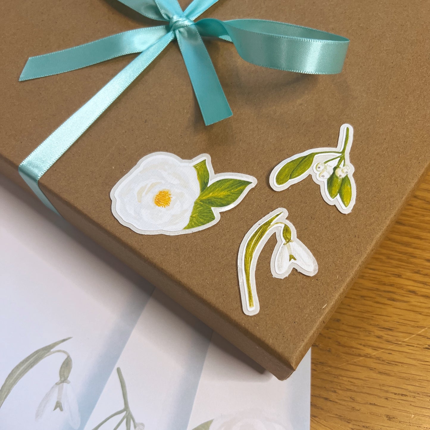 A snowdrop, white rose and mistletoe die cut stickers with easier to peel edge on a kraft presentation box wrapped in sea green ribbon