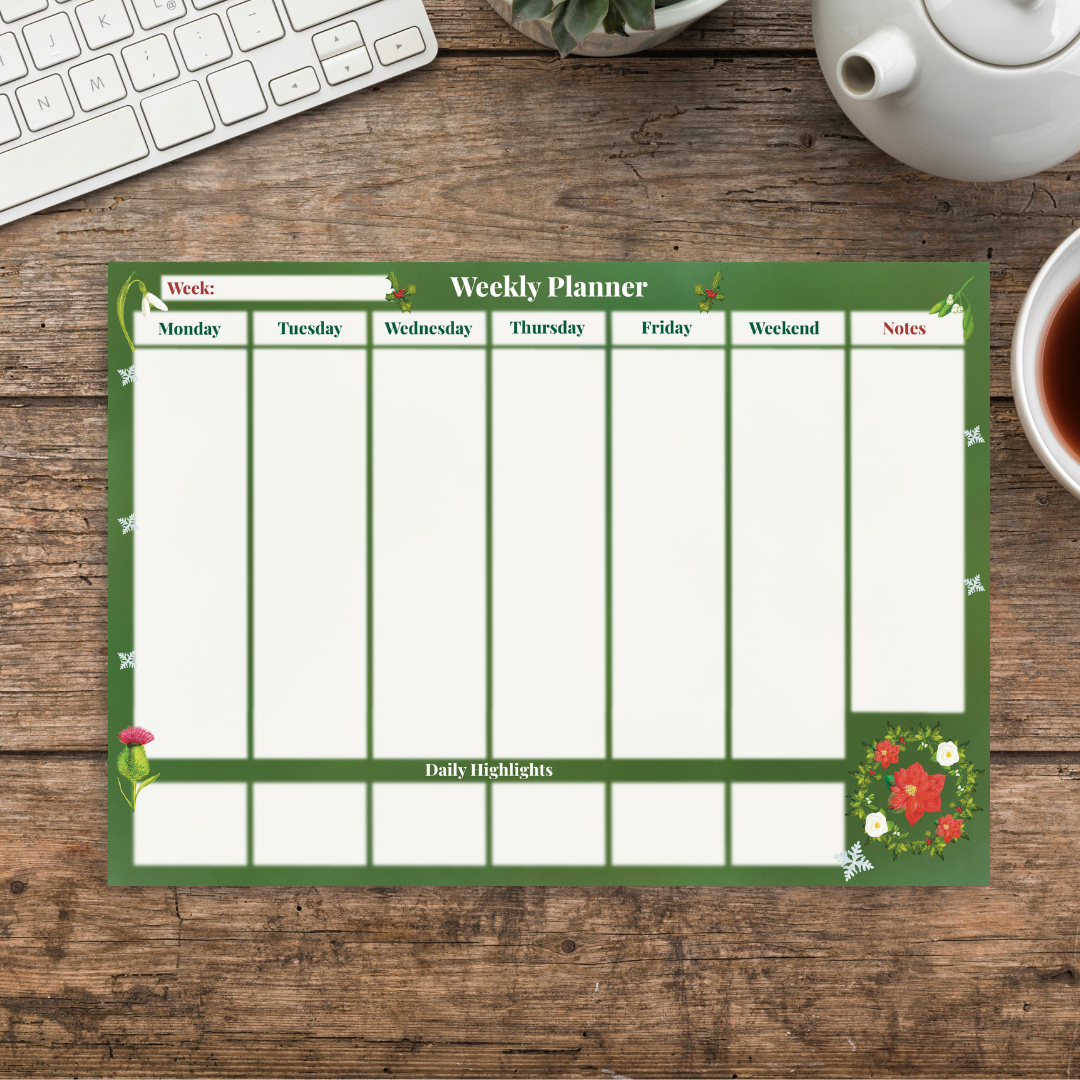 Winter botanical weekly planner with a festive green background, illustrated with winter florals and snowflakes.  White column sections for each day of the week have smaller daily highlights sections underneath each day, and to the left is a notes column for writing in also.  The pad sits on a wooden desk next to a cup of tea and a keyboard.