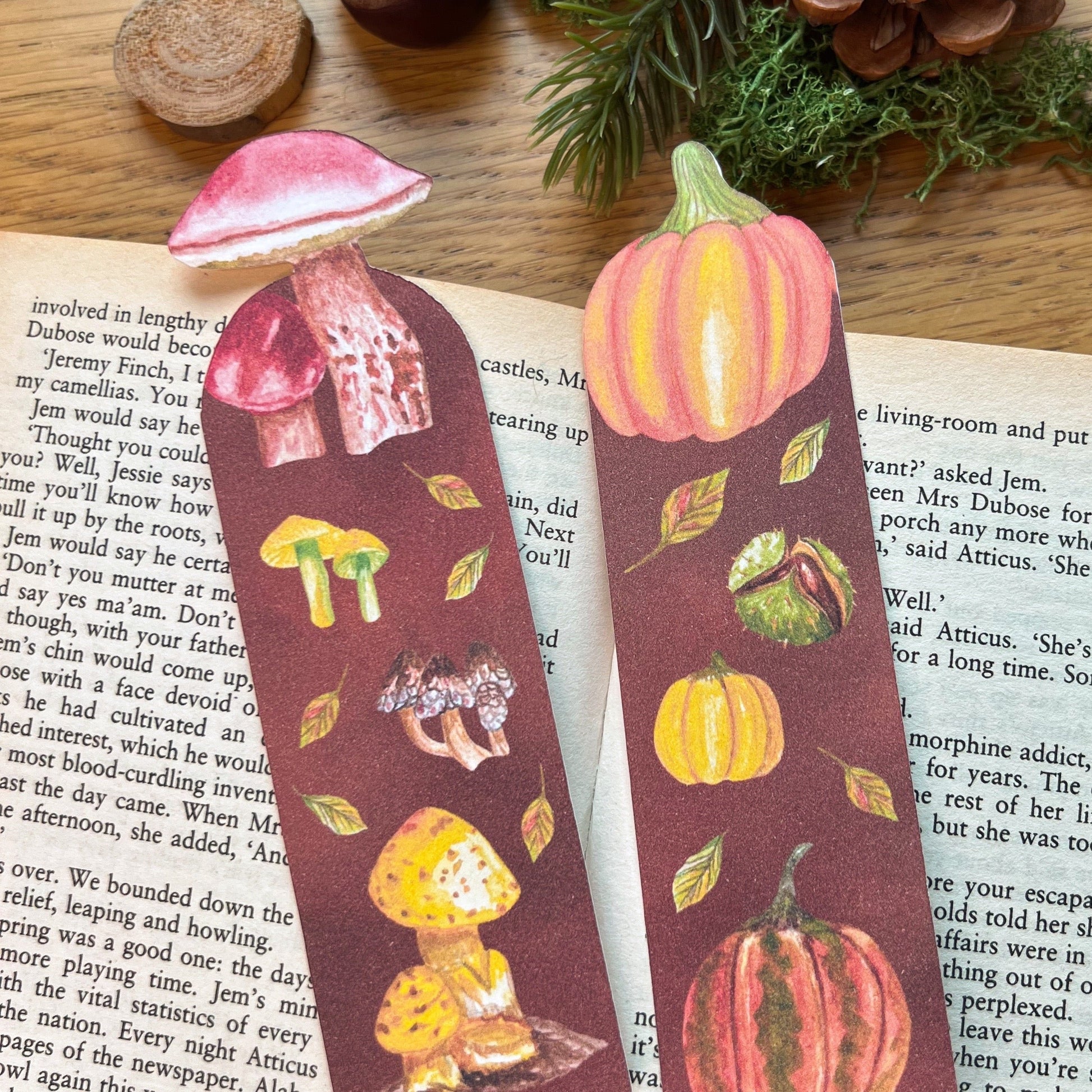 Fall Forage Pumpkin Bookmark featuring two orange and one yellow pumpkins with autmnal leabes tumbling dow the illustrated bookmark. An orange Cinderella pumpkin is cut out at the top, and the bookmark is resting on the open pages of a book next to the Fall Forage Mushrooms bookmark which features Mushrooms acorss the warm tones instead.